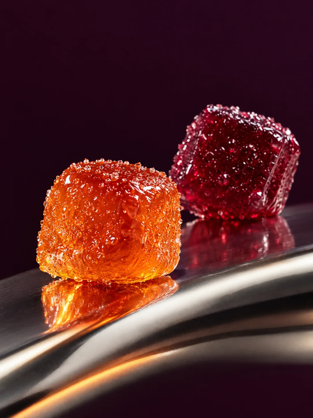 Blood-orange and berry Morsel gummies on curved chrome against deep aubergine