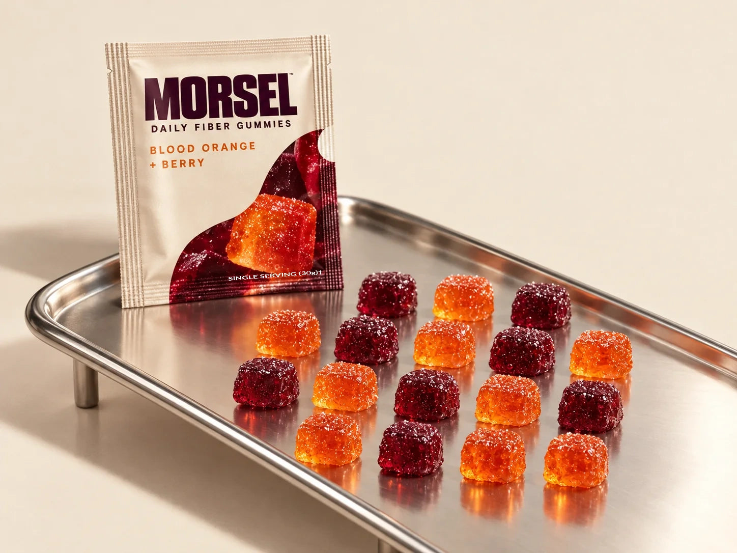 A cream Morsel pouch beside rows of blood orange and berry gummies on a polished steel tray